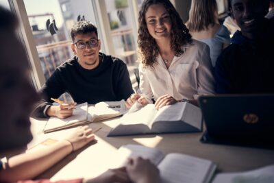 Studenter studerar tillsammans vid ett bord, omgivna av böcker och en bärbar dator